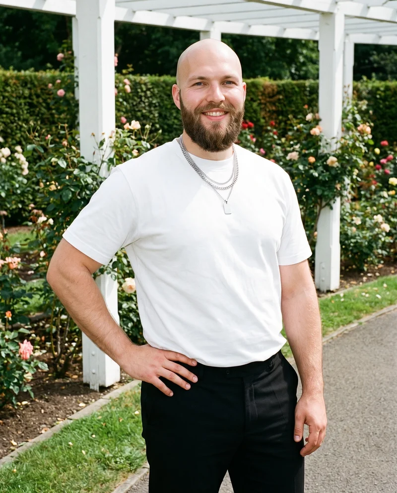 A 26-year-old Caucasian man wears a men's silver layered necklace in an upper-body, angled-front view. He is smiling with one hand on his hip, against a rose garden pergola backdrop.