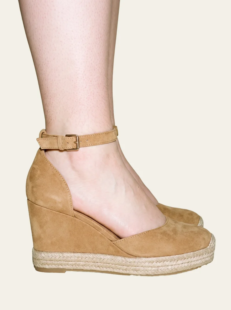 Tan espadrille wedges shown from the side on a flexed ankle, framed from the ankle down against an alabaster flat-color backdrop.