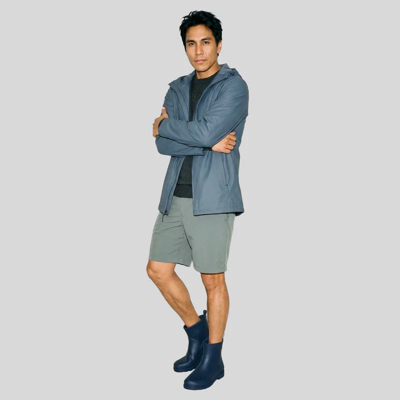 A 42-year-old Indigenous American man is shown full body from a front three-quarter angle wearing men's navy rubber rain boots. He stands in a self-hug pose with a natural facial expression against a flat pearl grey backdrop.