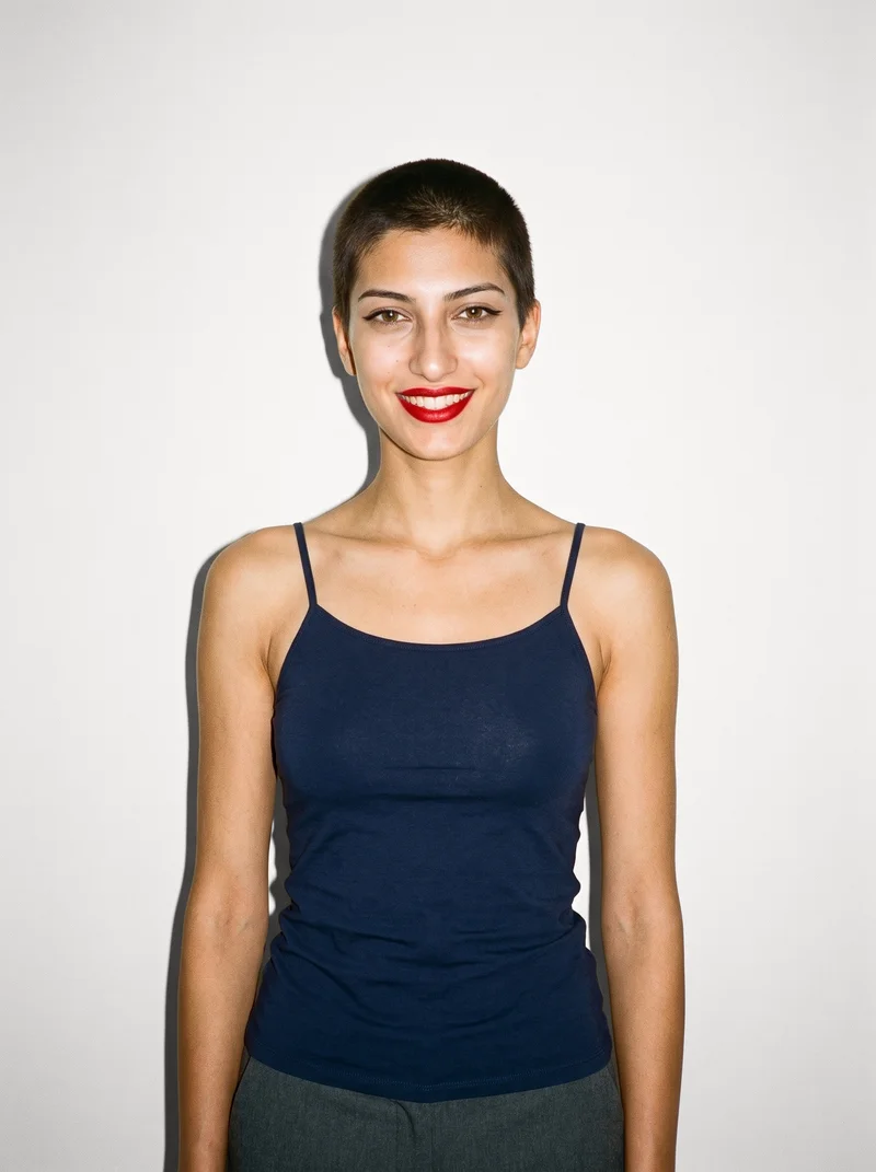 A 21-year-old Turkish woman wears a navy cami top in an upper-body, front-view photograph. Her arms are relaxed and she is smiling, with a Chantilly Lace studio-wall backdrop.