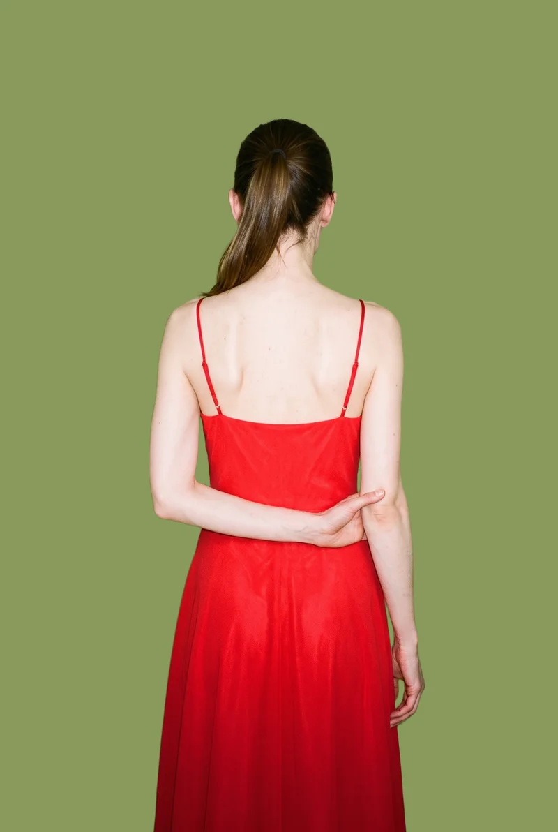 A 38-year-old Slavic woman wears a red chiffon maxi dress, shown from the back in a three-quarter-length view with one elbow positioned behind her back. A flat moss-green backdrop surrounds the figure.