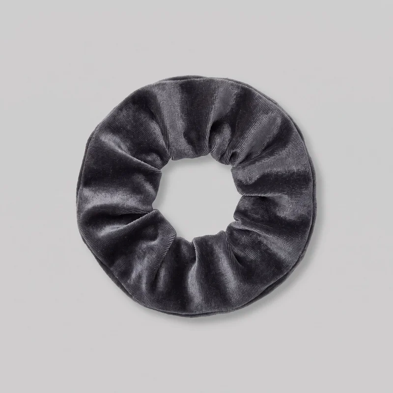 A complete velvet hair tie is shown laid flat from above as a gathered circular ring with a central opening. Its padded, fabric-covered elastic tube forms soft radial folds and pleats around the circumference, with rounded edges and subtle stitched seams along the perimeter. No closures, hardware, logos, prints, patches, embroidery, or embellishments are visible.