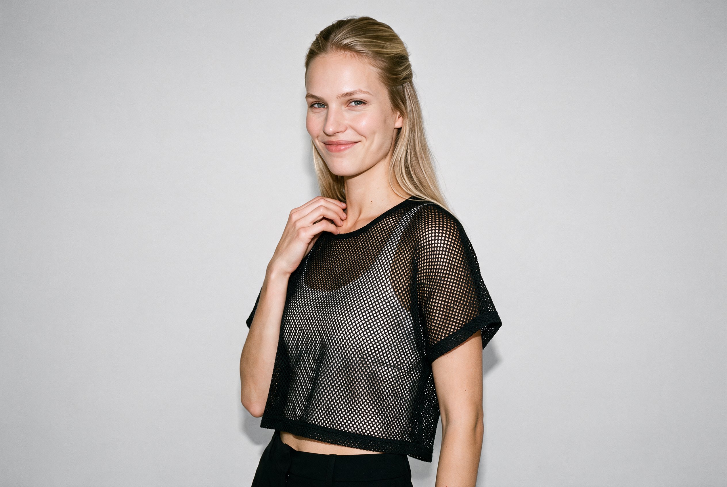 A 20-year-old Caucasian woman is shown from the upper body at an angled front view, wearing a women's black mesh cover-up. She is smiling with one hand resting at her collarbone. A fog-grey studio-wall backdrop fills the background.