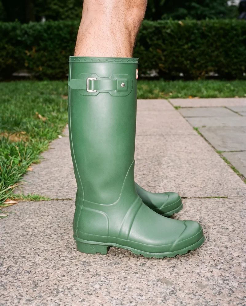 A men's green Wellington boot shown from the side at ankle level, framed around the ankle. A park terrace fountain forms the backdrop.