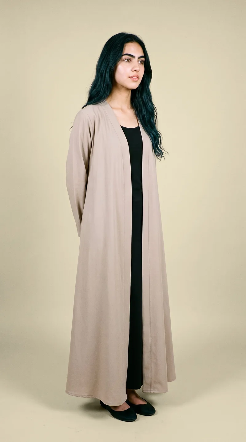 An 18-year-old Arab woman wears a beige open-front abaya in a full-body, angled-front view. She stands with her hands behind her back and a neutral facial expression against a flat wheat-colored backdrop.