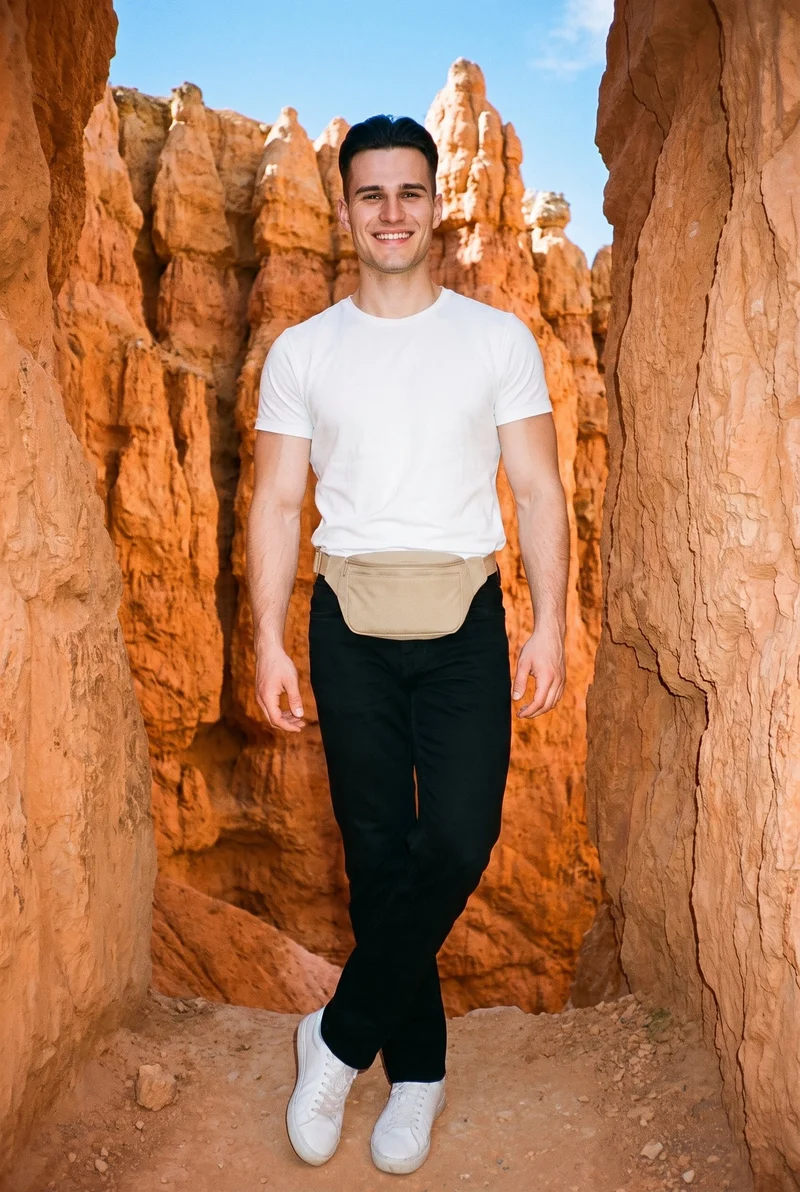 A 20-year-old Slavic man is shown full body from the front, wearing a men's beige belt bag. He stands with his ankles crossed and a grin, with Bryce Canyon hoodoos as the backdrop.