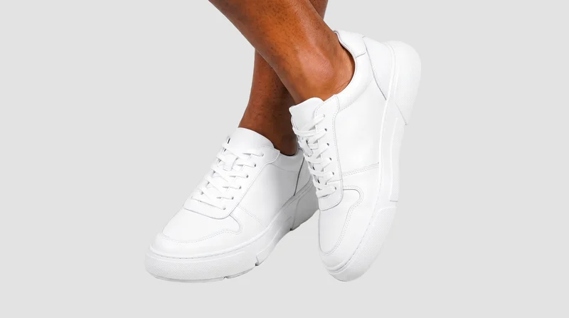 A pair of men's chunky white sneakers is shown from a front three-quarter angle with the ankles crossed. The shoes are framed against a flat fog-grey backdrop.