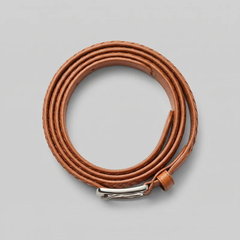 A complete tan woven leather belt is shown coiled into a circular shape as a standalone product. The braided strap has raised smooth edges, visible edge stitching around the fixed keeper loop, repeated diagonal openings from the woven construction, and multiple adjustment holes along the inner side. A curved metal buckle with a prong-style fastening is visible at one end.