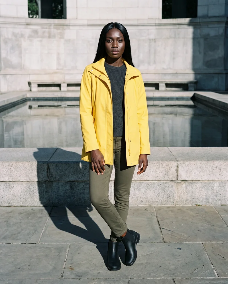 A 28-year-old African woman wears a yellow rain mac in a front view, posing with one hip angled outward and a confident expression. The Memorial Plaza serves as the environmental backdrop.