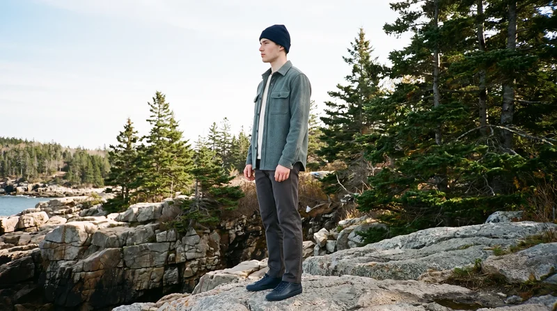 A 19-year-old Caucasian man wears a navy beanie in side view while standing in a relaxed pose with an intense expression. Acadia coastal rocks form the environmental backdrop.