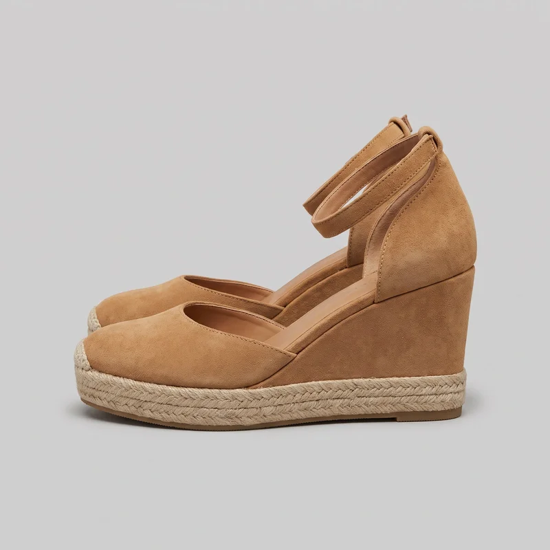 A pair of tan closed-toe wedge espadrille sandals is shown in a side-angled standalone view. The shoes have rounded toes, open side cutouts, covered heel counters, wraparound ankle straps, high wedge heels, and raised platforms edged with braided espadrille detailing.