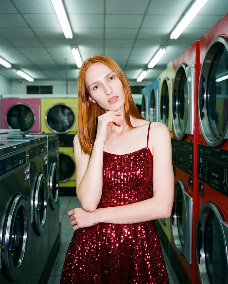 A 19-year-old Caucasian woman is shown from the upper body in a red sequin cocktail dress, facing forward with one hand at her chin and a natural expression. A laundromat backdrop is visible behind her.
