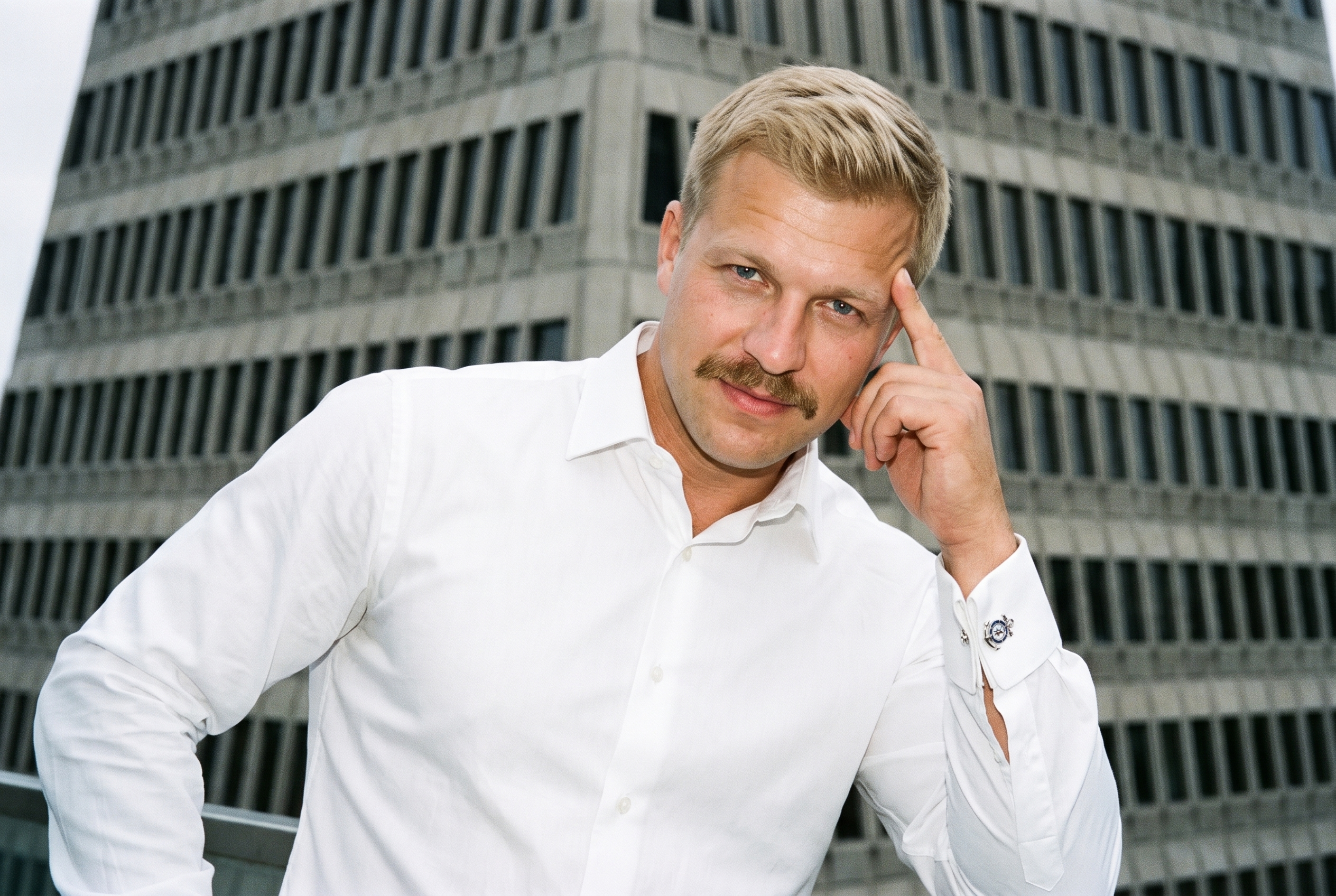 A 41-year-old Slavic man is shown in an upper-body frame from a front three-quarter angle, wearing themed novelty cufflinks. He poses with one hand at his temple and a confident facial expression. A low-angle pyramid-building backdrop is visible behind him.