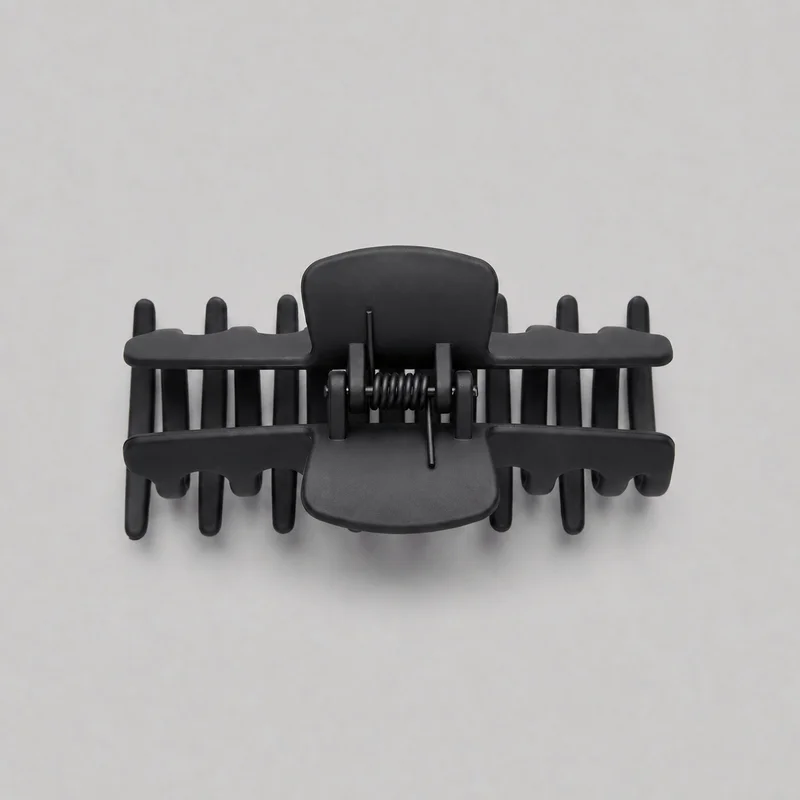 A mostly complete black claw hair clip is shown as a standalone product from an above-front angle. Its hinged clamp has two opposing comb sections joined by a central coiled spring and pivot hardware, with rounded rectangular press tabs, an arched central back plate, parallel teeth, scalloped gripping edges, and rounded outer corners. The smooth exterior is unadorned, with no visible logos or embellishments.