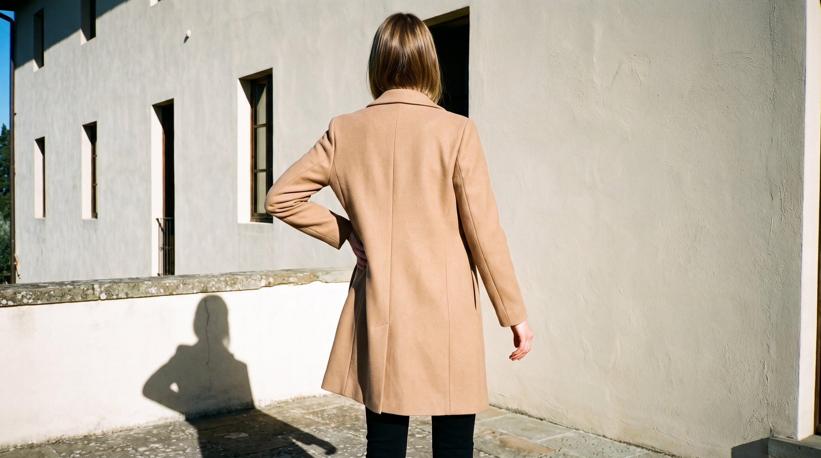 A 24-year-old Caucasian woman wears a camel wool coat in a three-quarter-length rear view, with one hand on her hip. An Italian villa terrace forms the backdrop.
