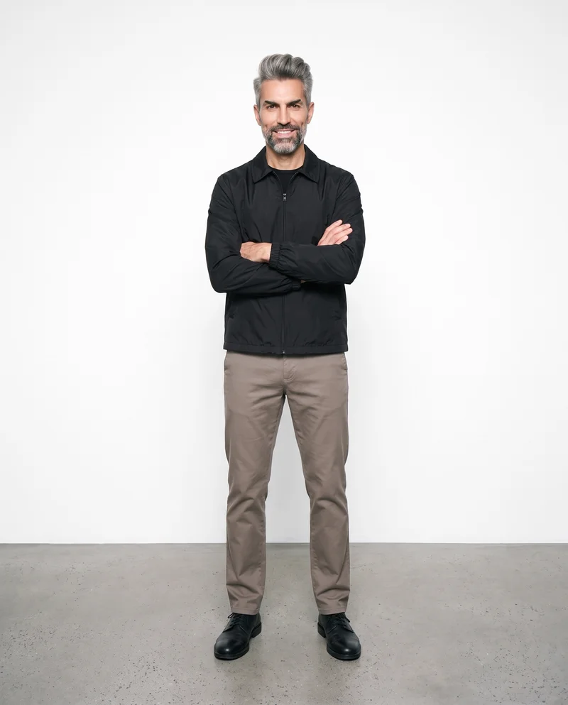 A 47-year-old Hispanic man wears a men's black windbreaker in a front-facing environmental composition. He is smiling and holding himself in a self-hug pose against a pure white studio-wall backdrop.