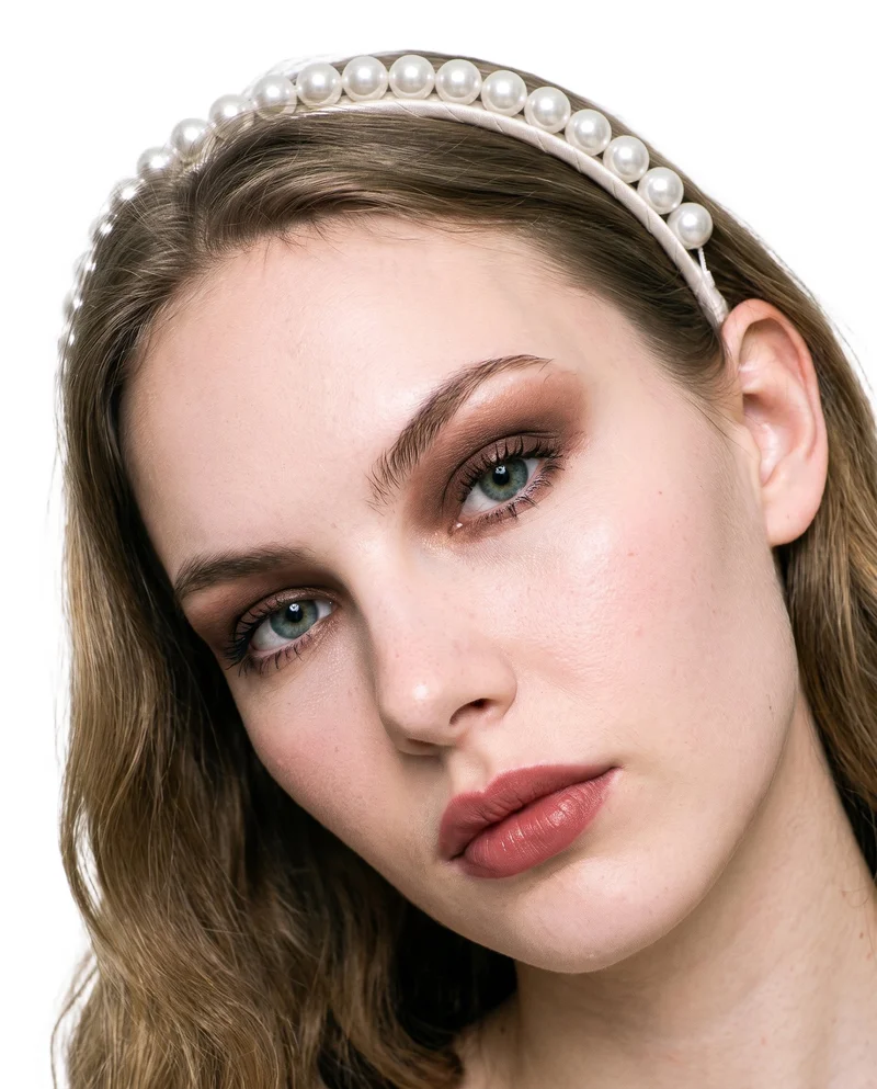 An 18-year-old Caucasian female model wears a pearl headband in a front-view head shot. Her head is tilted and her expression is neutral, with a pure white flat-color backdrop.
