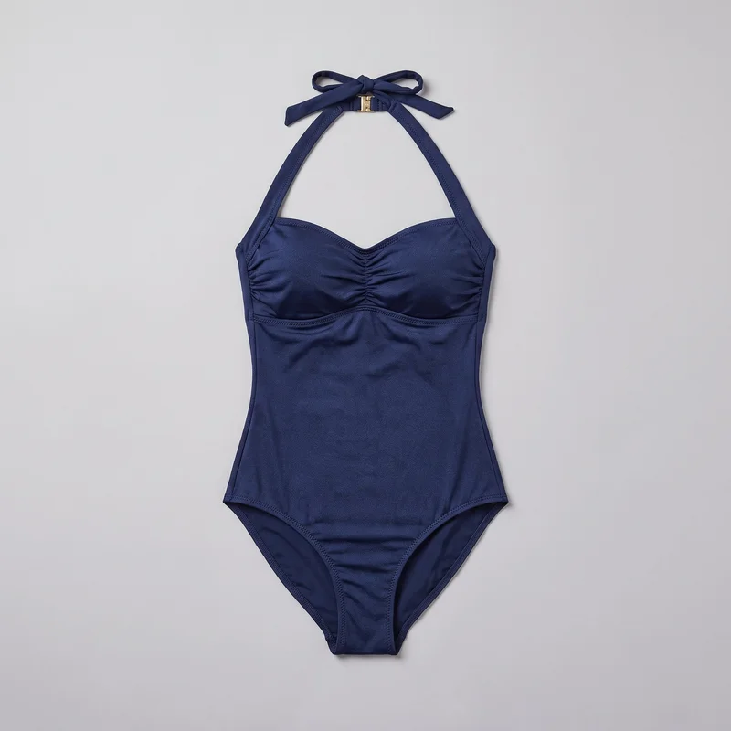 A navy women's one-piece halter swimsuit is shown in a complete flat-lay front view. It has wide straps tied in a neck bow, a sweetheart neckline with a center notch, a small hardware accent below the bow, ruched gathering at the center bust, curved underbust seams, shaped side seams, bound edges, and high-cut leg openings.