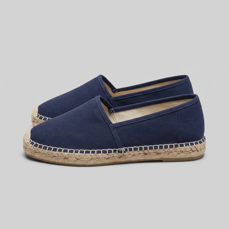 A pair of women's navy canvas espadrille flats is shown as a standalone product from a side and slight three-quarter angle. The low-profile slip-ons have closed rounded toes, low-cut openings, plain textile uppers, center seams, bound edges, visible topline stitching, and small elastic insets near the insteps. Braided rope wraps around the midsoles and toe edges, with contrasting loop stitching along the upper-to-sole seams and thin flat outsoles beneath.