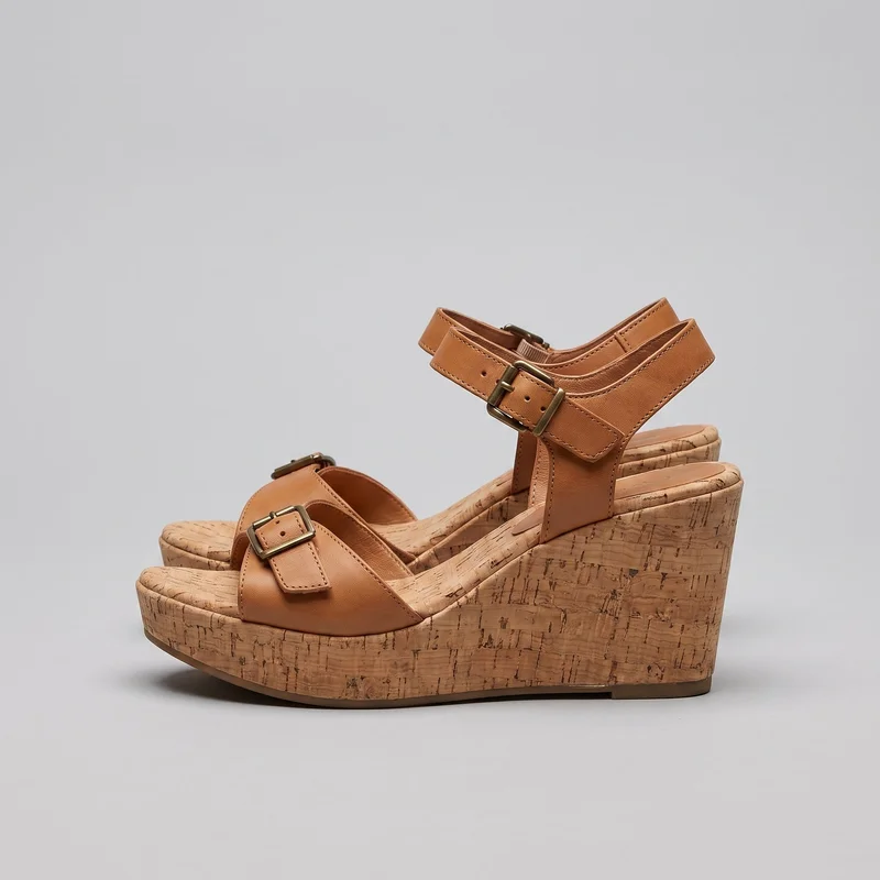 A pair of women's open-toe platform wedge sandals is shown from a side three-quarter angle. The sandals have broad forefoot vamp straps and wide ankle straps with rectangular buckles, punched adjustment holes, and stitched edging, along with vertical side and heel straps. Their contoured footbeds, rounded open toes, raised forefoot platforms, high wedge heels, cork-grain exterior pattern, and thin outsole edges are visible.