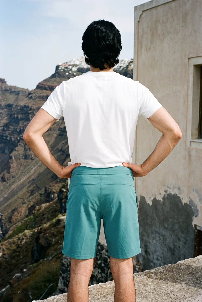 A 19-year-old Caucasian male model is shown from the rear in a three-quarter-length view, wearing teal board shorts with his hands on his hips. A Santorini caldera view forms the backdrop.