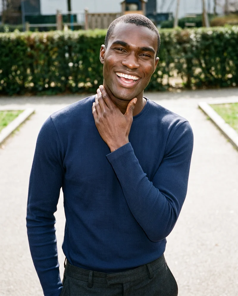 A 25-year-old African man wears a navy long-sleeve thermal top in an upper-body front view. He is laughing with one hand on his neck, against a playground backdrop.