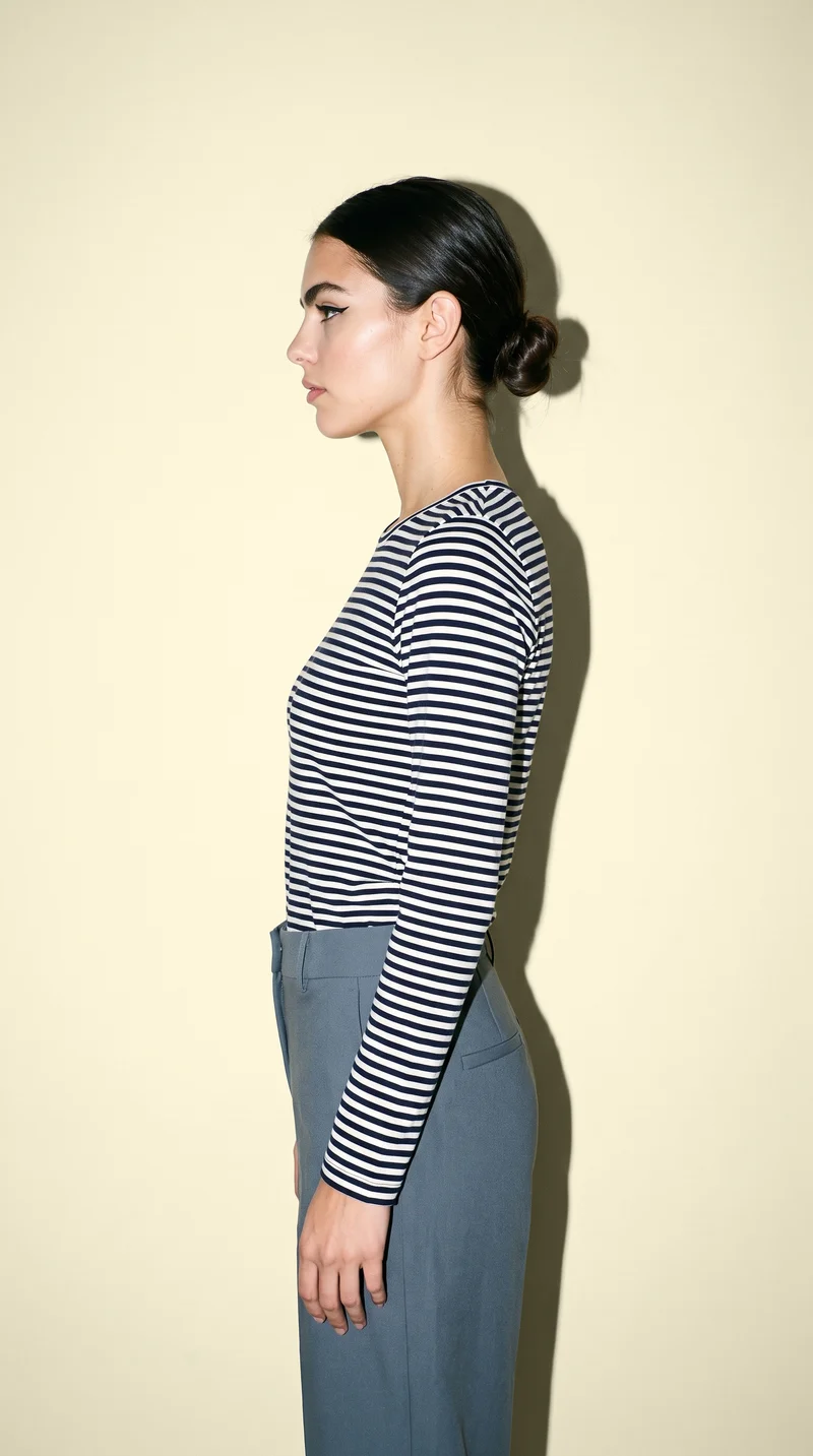 A 22-year-old Caucasian woman models a women's navy striped long-sleeve tee in a three-quarter-length side view, with her waist twisted and a serious expression. A buttercream studio-wall backdrop frames the composition.