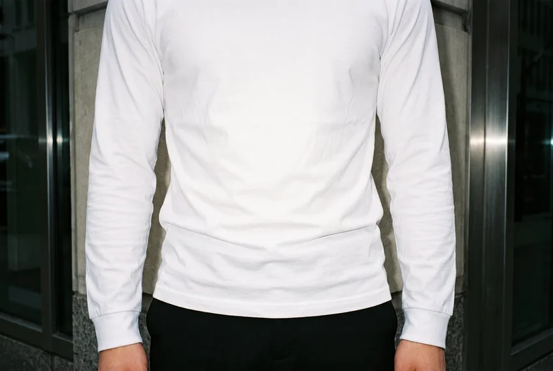 A man wears a white long-sleeve T-shirt in a front-facing, hips-level view cropped at the hips. A Financial District urban canyon forms the backdrop.
