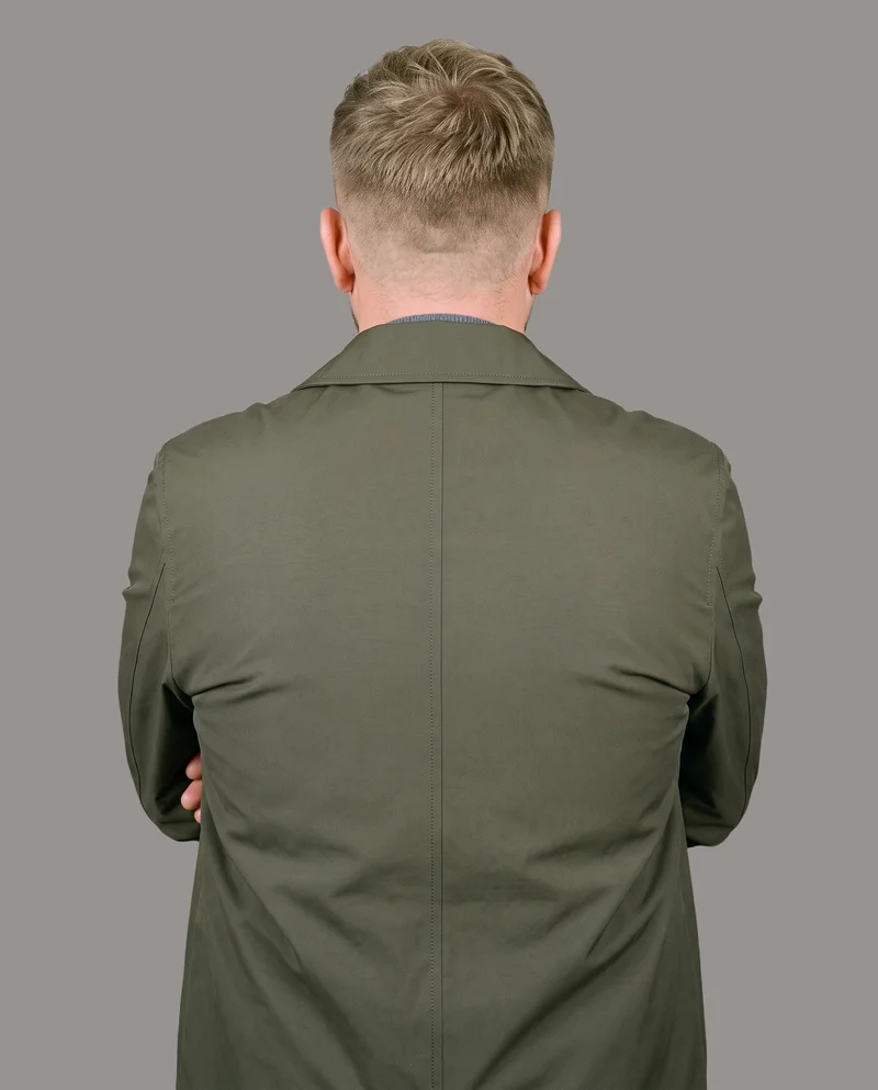 A 54-year-old Slavic man is shown from the upper body in a back view wearing an olive packable raincoat, with his arms crossed. The garment is framed against a flat studio grey backdrop.