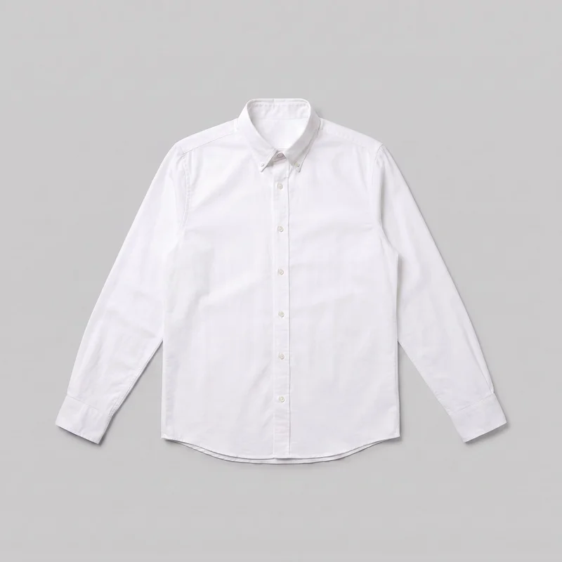 A white Oxford shirt is laid flat in a mostly complete front view as a standalone product. It has a pointed button-down collar, full-length front button placket, long set-in sleeves with structured cuffs, visible shoulder and yoke seams, plain front panels without chest pockets, and a curved shirttail hem with a layered back edge visible below the front.