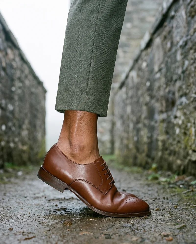 Brown derby shoes shown from the side while positioned on tiptoe, framed from the ankles against a Scottish castle backdrop.
