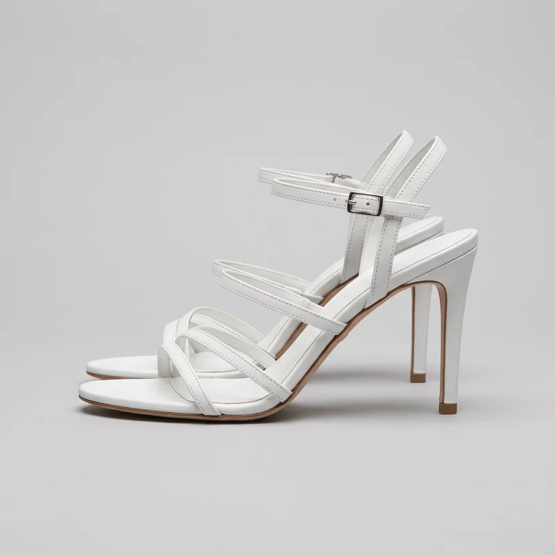A pair of white strappy high-heel sandals is shown in a side-oriented standalone view. The sandals have open toes, multiple narrow straps crossing the forefoot, ankle straps with rectangular buckle closures, thin soles, and tall straight stiletto heels.