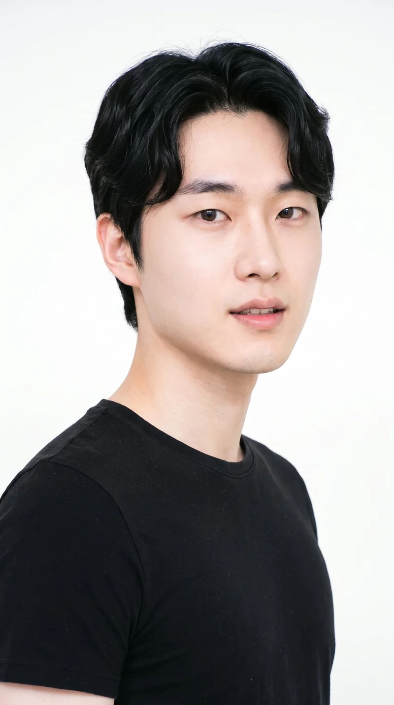 This 27-year-old Korean man has light porcelain skin with a neutral undertone and East Asian facial features. His face is round, with soft, full contours, gentle curves, and few angles. He has medium-thickness eyebrows, very dark near-black eyes, and medium lips. His deep black hair falls in soft waves with gentle movement. He is clean-shaven, with a smooth face and no facial hair.