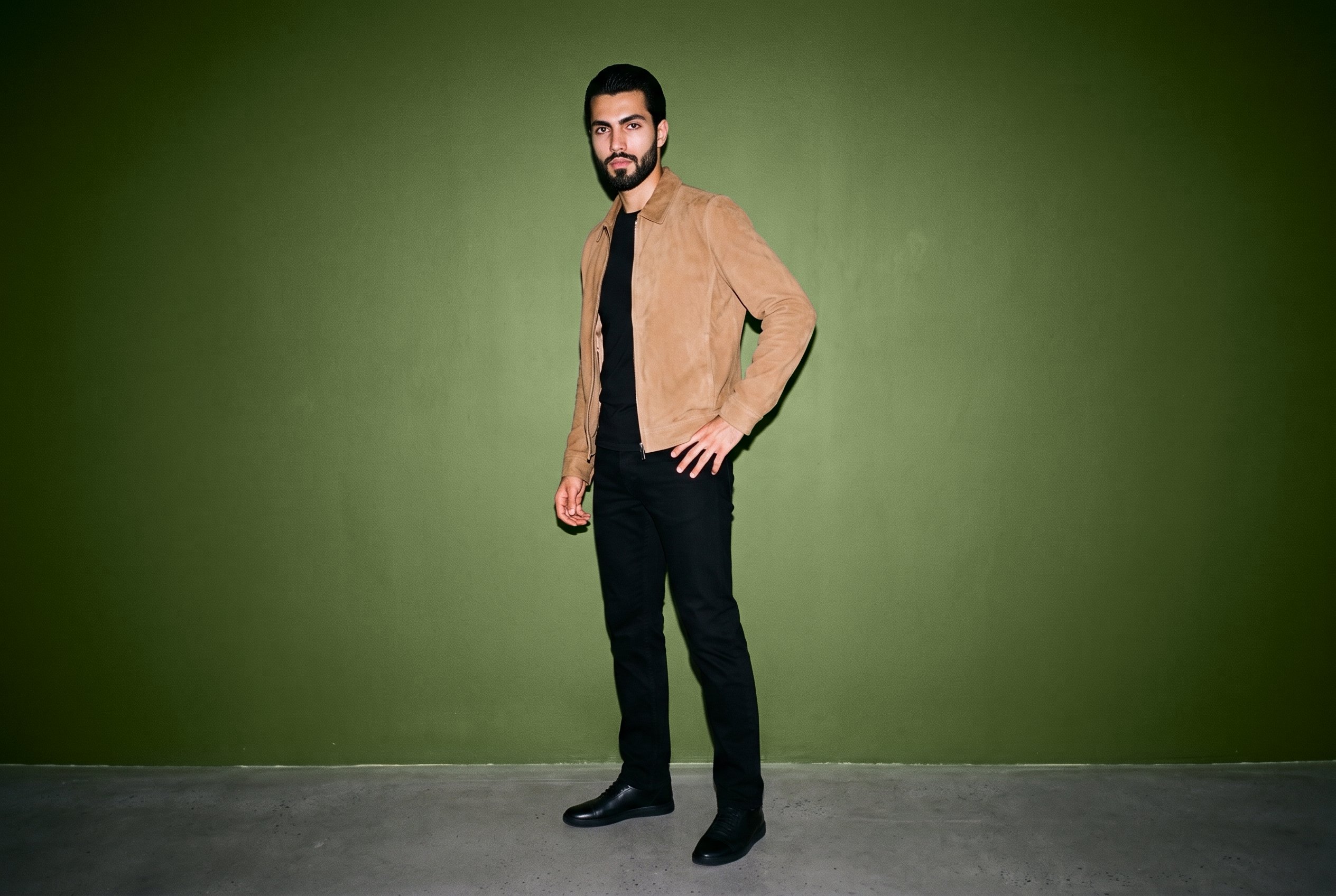A 23-year-old Caucasian man wears a men's tan suede jacket in a full-body angled-front view, posing with one hand on his hip and an editorial expression. An olive grove studio-wall backdrop frames the scene.