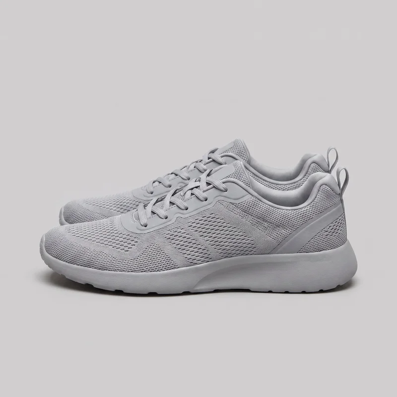 A pair of women's grey mesh trainers is shown in a side-oriented standalone view. The low-top athletic sneakers have rounded toes, ventilated grid-textured uppers with oval openings, lace-up fronts threaded through molded eyestay panels, padded collars and tongues, heel pull loops, diagonal side overlays, and thick continuous cushioned soles with shallow tread visible along the bottom edge.