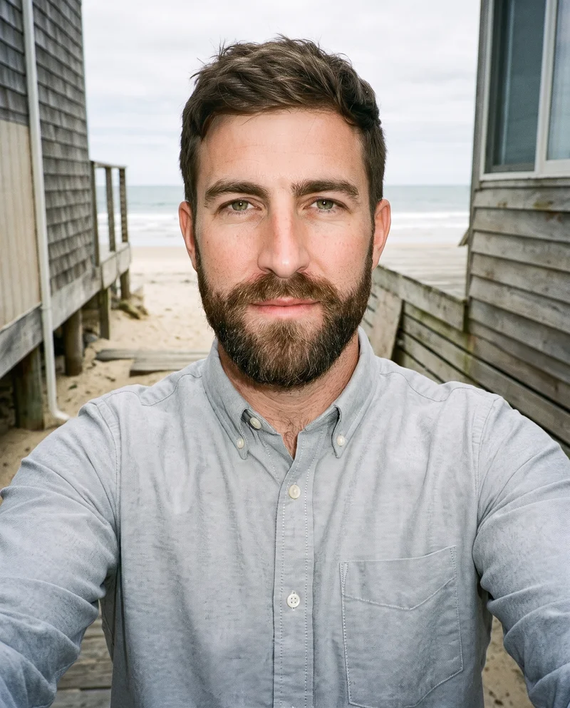 A 36-year-old Caucasian man wears a grey cotton Oxford shirt in a front-view selfie. His chin is lowered with a natural facial expression, against a Cape Cod Beach House backdrop.