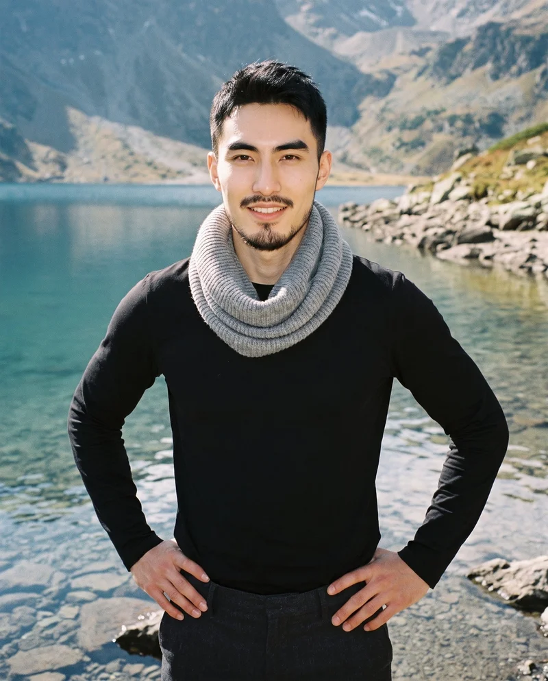 A 25-year-old Chinese man wears a grey knit snood in an upper-body front view, standing with his hands on his hips and smiling. An alpine lake shore forms the backdrop.