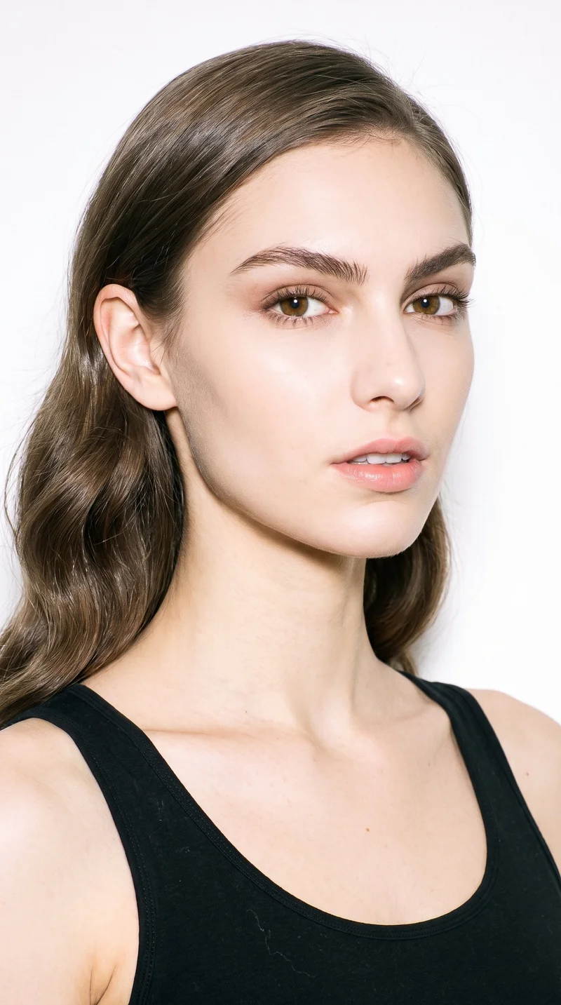 A 20-year-old Caucasian woman with porcelain skin and a warm golden undertone. Her diamond-shaped face has a narrow forehead and jaw, with the greatest width at the cheekbones. She has thick eyebrows, brown eyes, and thin lips. Her ash-brown hair has a cool undertone and is styled in large, polished Hollywood waves.