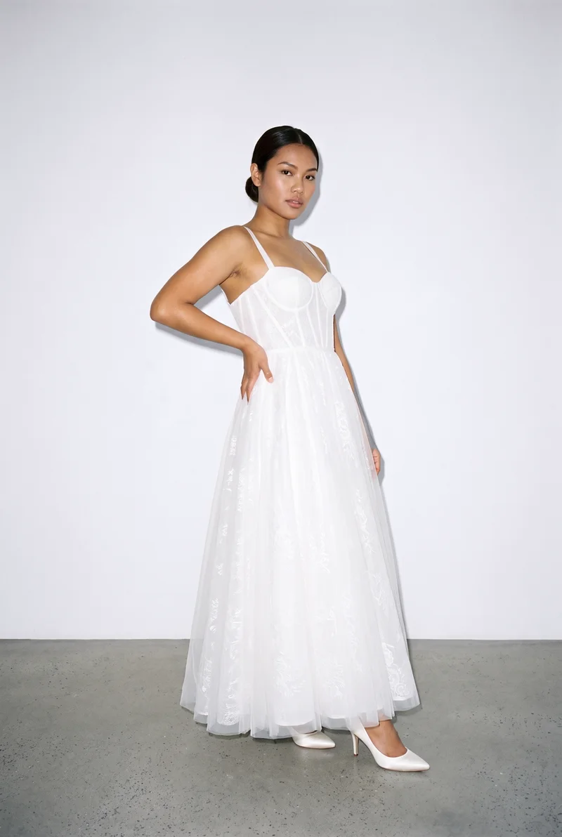 A 23-year-old Southeast Asian woman wears a white A-line wedding dress in a full-body view from a front three-quarter angle. She stands with her weight back and a neutral facial expression against a porcelain studio-wall backdrop.
