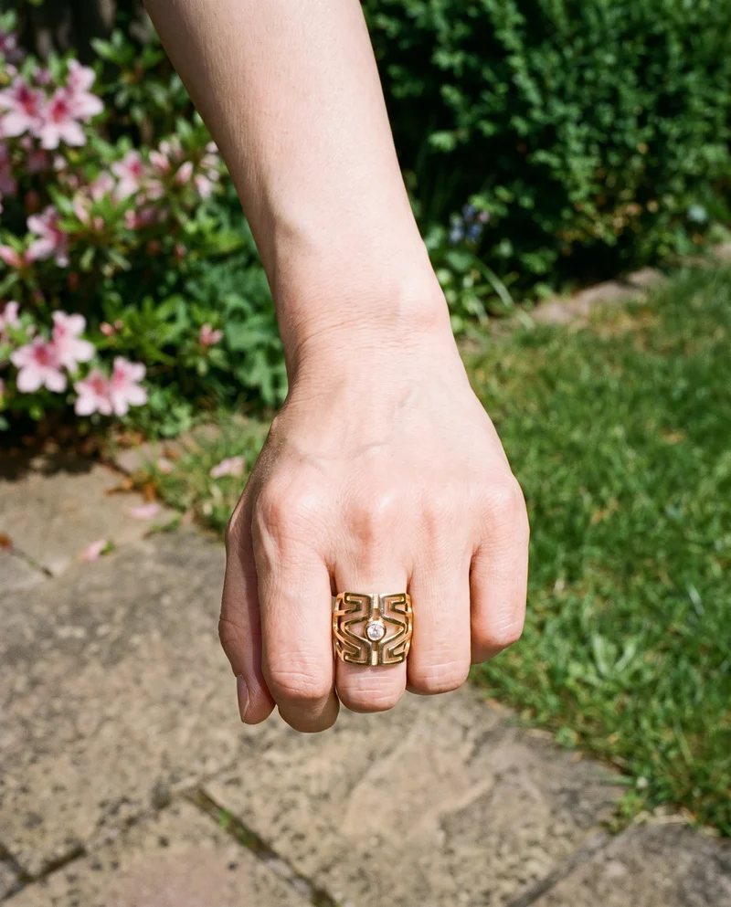 A hand and wrist wearing a gold statement ring are shown from above with the knuckles facing forward. An Azalea Garden backdrop is visible.