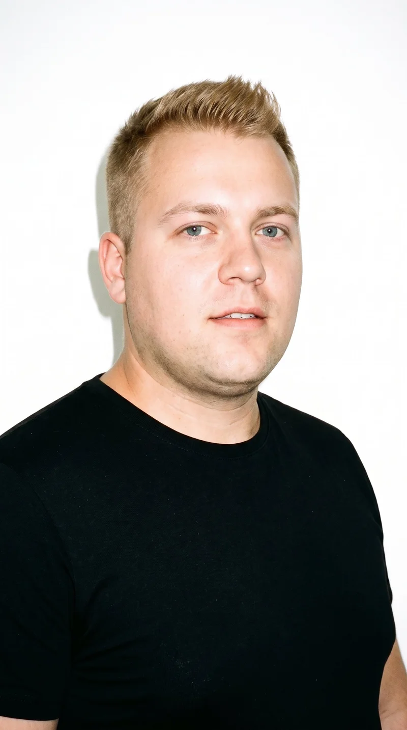 A 33-year-old Caucasian man with medium olive skin and a peach undertone has a round face with soft, full contours and few angles. His thin eyebrows frame cool, clear grey eyes, and his medium lips sit above a clean-shaven, smooth face. His darker, slightly muted blonde hair is styled in a short crew cut, tapered with the sides slightly shorter than the top.