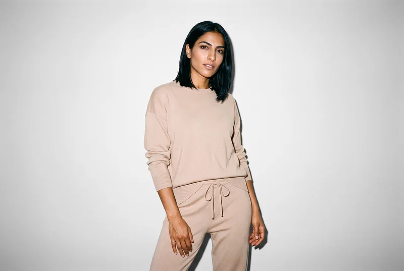 A 34-year-old Arab woman wears a beige knit loungewear set against a fog grey studio-wall backdrop. She is shown from the front in a three-quarter-length frame with a waist-twist pose and a natural facial expression.
