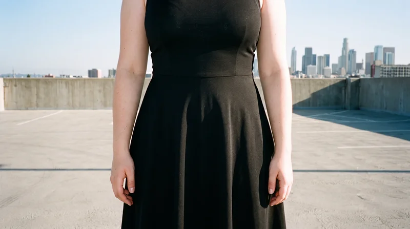 A woman models a black midi dress in a front-facing, waist-level composition. The parking garage rooftop serves as the backdrop.
