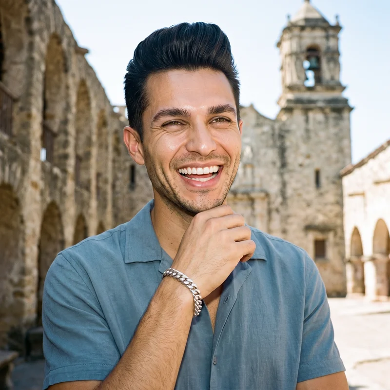 A 45-year-old Hispanic man is shown in an upper-body front view wearing a men's Cuban link bracelet, with one hand at his chin as he laughs. The Mission Courtyard Bell Fountain appears as the backdrop.