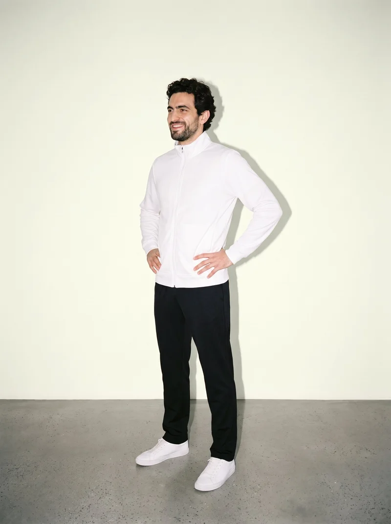 A 33-year-old Caucasian man wears a white track jacket in a full-body, angled-front view with his hands on his hips and a natural expression. An ivory lace studio-wall backdrop is visible behind him.
