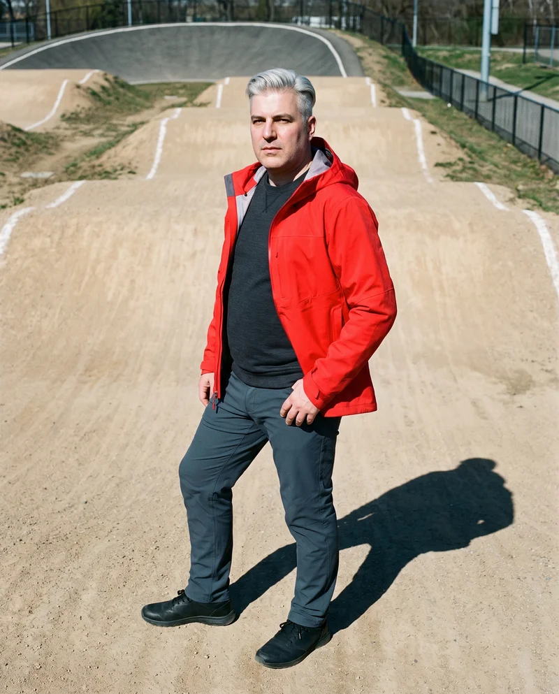A 51-year-old Caucasian man wears a red hardshell jacket in a 3/4-length side view. He stands with his weight back and a neutral facial expression, with a BMX track backdrop.