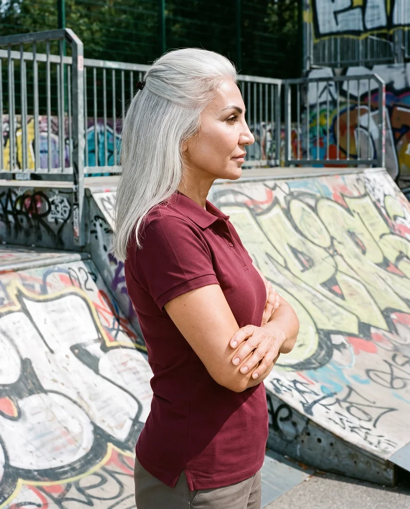 A 58-year-old Turkish woman is shown from the upper body in side view wearing a burgundy pique polo shirt, with her arms crossed and a natural expression. A skate park serves as the backdrop.