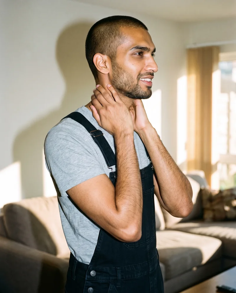 A 34-year-old Indian man wearing black denim dungarees is shown from the side in an upper-body view, smiling with his hands on his neck. A home living room forms the backdrop.
