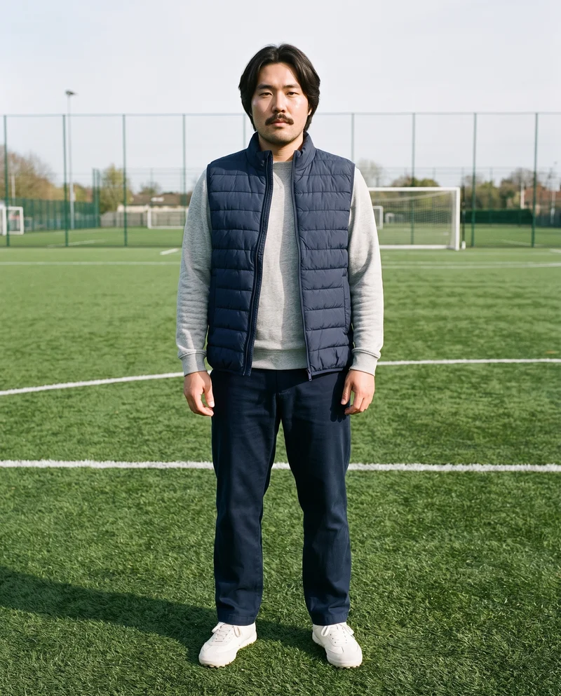 A 32-year-old Chinese man is shown full body from the front wearing a navy padded gilet. He stands with his ankles crossed and a fierce facial expression, against a soccer pitch backdrop.