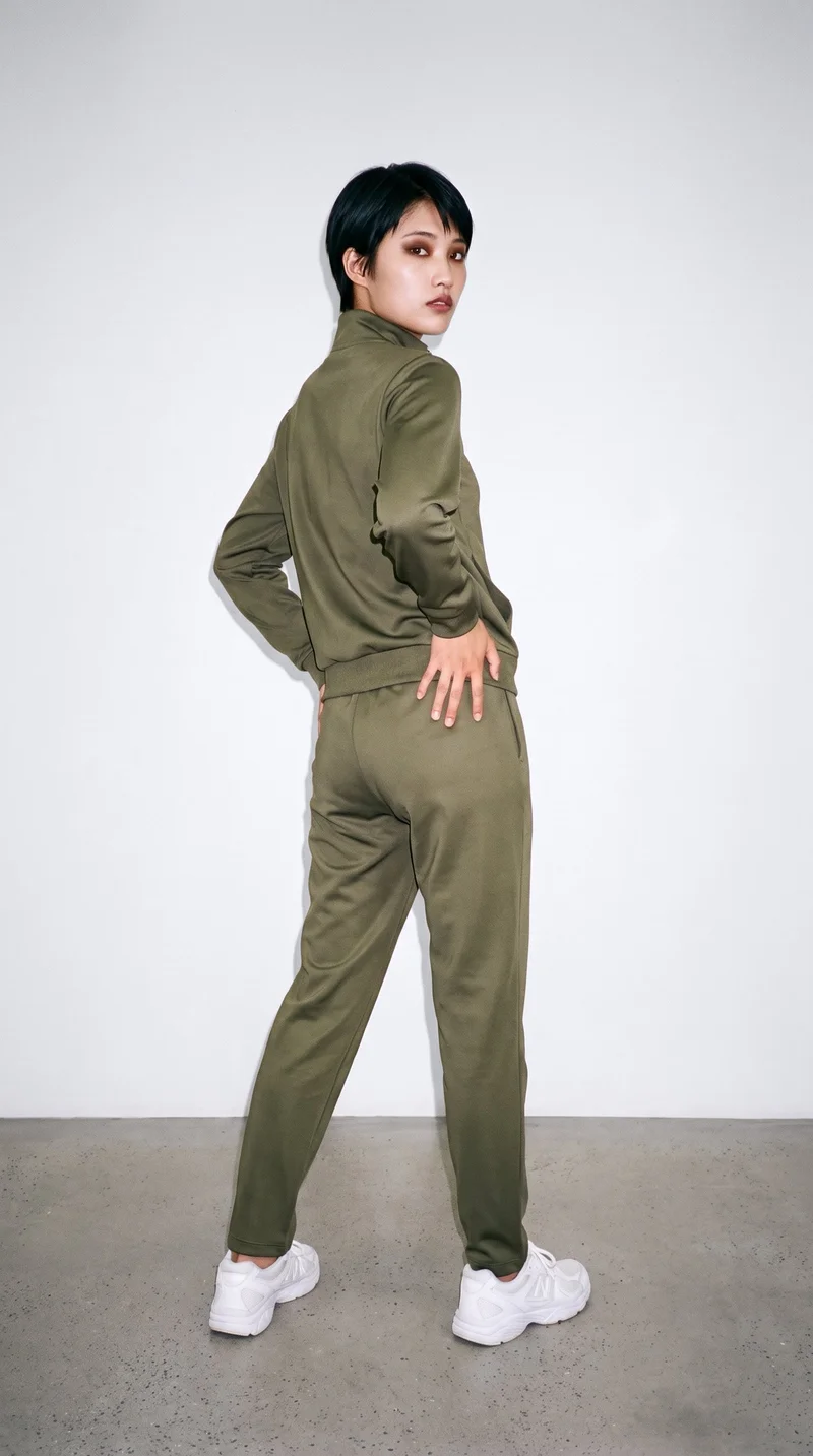 A 19-year-old Japanese woman wears a women's olive tracksuit in a full-body view from an angle behind, with one hand on her hip. A porcelain studio-wall backdrop frames the composition.